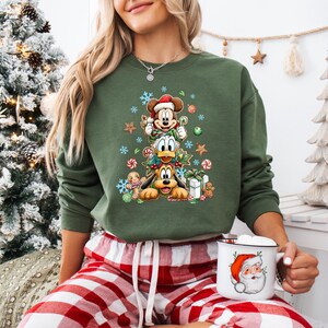 Peut inclure: Sweat-shirt vert avec Mickey Mouse, Donald Duck et Pluto dispos&eacute;s en sapin de No&euml;l, orn&eacute; de flocons de neige, de cannes en sucre et de bonhommes en pain d'&eacute;pices. Une tasse du P&egrave;re No&euml;l est tenue &agrave; la main. V&ecirc;tements de vacances festifs.