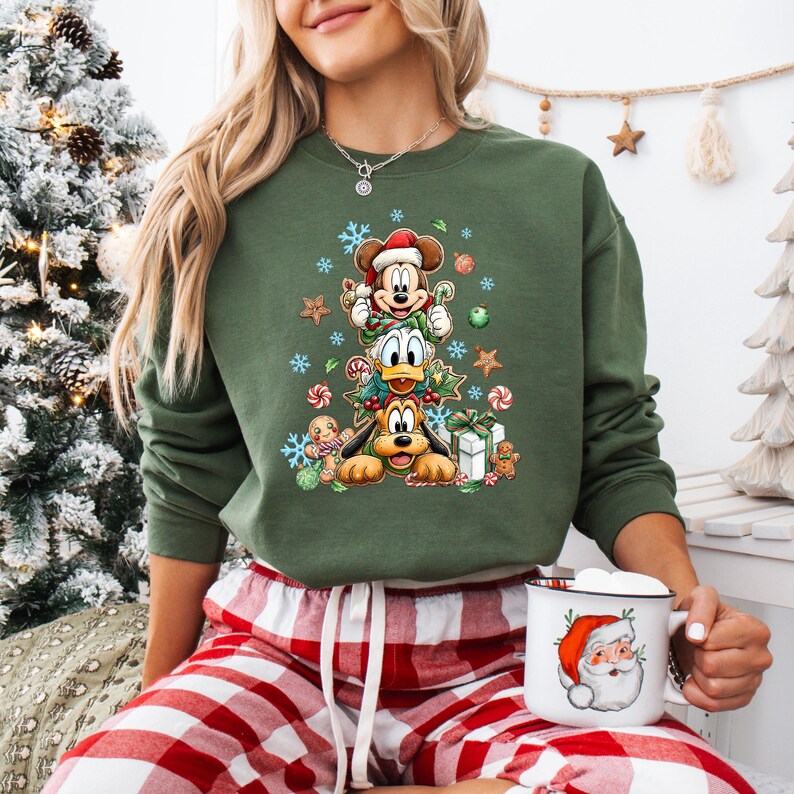 Peut inclure: Sweat-shirt vert avec Mickey Mouse, Donald Duck et Pluto dispos&eacute;s en sapin de No&euml;l, orn&eacute; de flocons de neige, de cannes en sucre et de bonhommes en pain d'&eacute;pices. Une tasse du P&egrave;re No&euml;l est tenue &agrave; la main. V&ecirc;tements de vacances festifs.