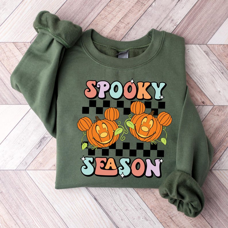May include: Green sweatshirt with the words "SPOOKY SEASON" in colorful retro font. The design features two smiling jack-o'-lanterns with mouse-ear-shaped pumpkins on a black and white checkered background. A fun Halloween-themed design.