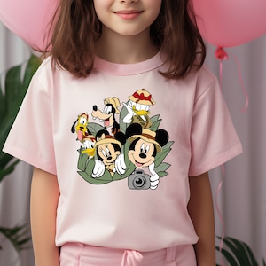 May include: Pink t-shirt featuring a graphic print of Mickey Mouse, Minnie Mouse, Goofy, Donald Duck, and Pluto wearing safari hats and holding a camera. The characters are surrounded by green leaves.