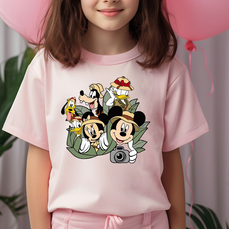 May include: Pink t-shirt featuring a graphic print of Mickey Mouse, Minnie Mouse, Goofy, Donald Duck, and Pluto wearing safari hats and holding a camera. The characters are surrounded by green leaves.