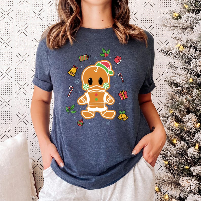 May include: A navy blue t-shirt with a gingerbread man wearing a Santa hat and a face mask. The gingerbread man is surrounded by Christmas-themed decorations, including candy canes, bells, and presents.