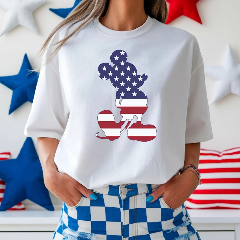 May include: White t-shirt with a Mickey Mouse silhouette design, filled with the American flag pattern. The design features blue stars on a navy background for the head and red and white stripes for the body and feet. The shirt is worn with blue and white checkered shorts.