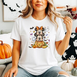 May include: White t-shirt with a Halloween graphic. The design shows Mickey and Minnie Mouse characters atop a carved pumpkin, with ghosts, bats, and sweets. A casual, festive apparel item, perfect for the spooky season.