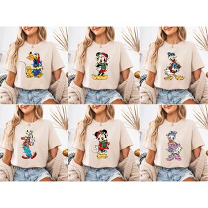 K&ouml;nnte beinhalten: Sechs beige T-Shirts mit Disney-Charakteren in Weihnachtskleidung. Micky Maus, Donald Duck und Goofy sind mit festlichen Lichtern und Accessoires dargestellt. Die Shirts werden auf einer Person pr&auml;sentiert.