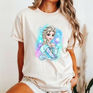 Op de afbeelding: Een licht beige T-shirt met een kleurrijke illustratie van Elsa uit Frozen. De illustratie toont Elsa met haar kenmerkende vlecht en blauwe jurk, omringd door sneeuwvlokken en een aquarel achtergrond.