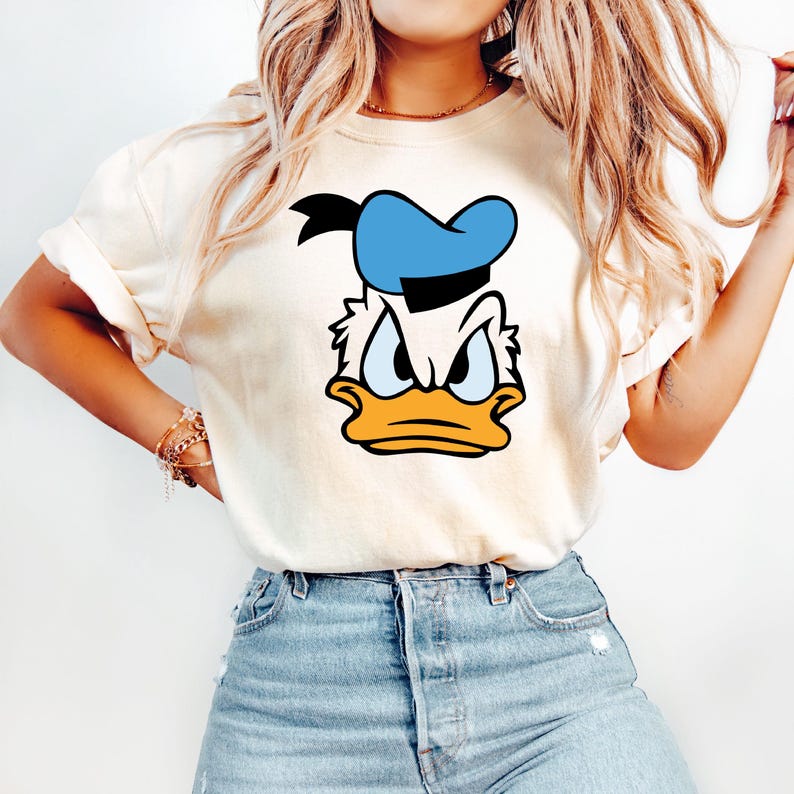 May include: Cream-colored t-shirt featuring a cartoon graphic of Donald Duck's face. The design includes a blue sailor hat, an orange beak, and an angry expression. The shirt is paired with blue denim jeans.