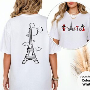 May include: White t-shirt featuring a black Eiffel Tower design with Mickey Mouse ears and clouds on the back. The front of the shirt has the word "PARIS" with Mickey and Minnie Mouse characters on either side. The shirt is a Comfort Colors brand.