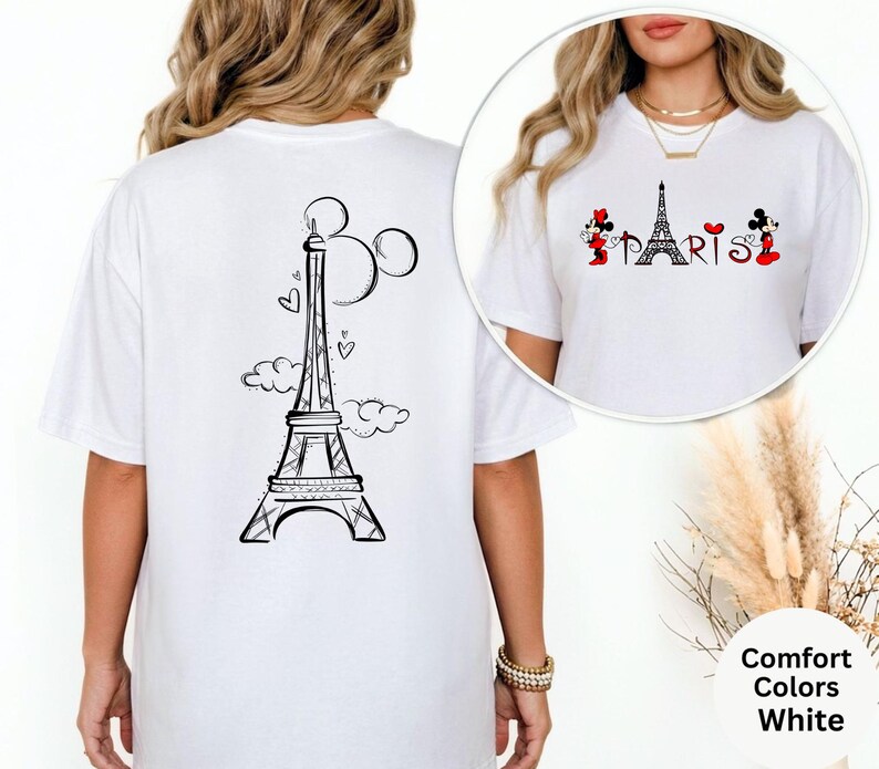 May include: White t-shirt featuring a black Eiffel Tower design with Mickey Mouse ears and clouds on the back. The front of the shirt has the word "PARIS" with Mickey and Minnie Mouse characters on either side. The shirt is a Comfort Colors brand.
