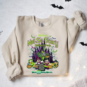 May include: A beige sweatshirt with a green, purple, and black graphic print of Mickey Mouse and friends in front of a castle. The text "Mickey's Not-So-Scary Halloween Party" is printed on the sweatshirt. The text "Walt Disney World" is printed at the bottom of the graphic.