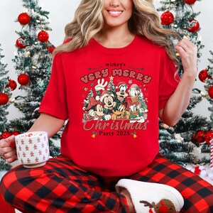 Puede incluir: Camiseta roja de manga corta con un dise&ntilde;o de Mickey Mouse y sus amigos, con el texto "mickey's Very Merry Christmas Party 2025". La persona lleva un pantal&oacute;n de pijama a cuadros rojos y negros. Se sostiene una taza blanca.