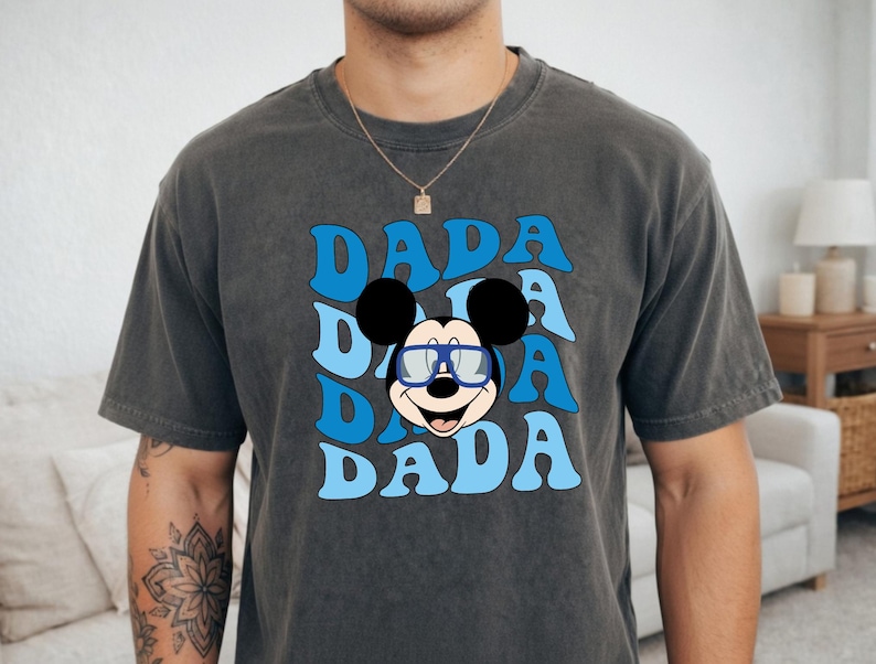 K&ouml;nnte beinhalten: Dunkelgraues T-Shirt mit dem Wort "DADA" in Blau, dreimal gestapelt, und einer Cartoon-Mickey-Mouse mit Brille. Das T-Shirt hat einen Vintage-Look.