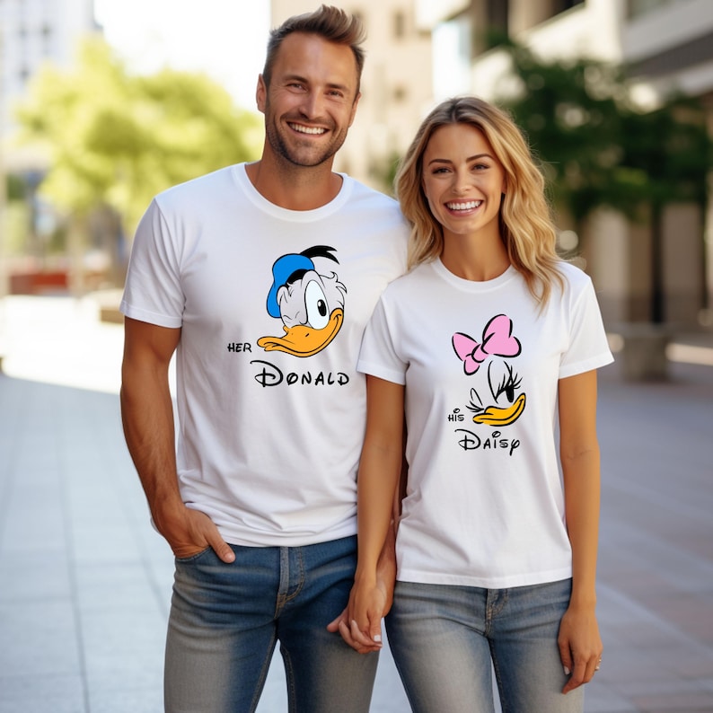 May include: White t-shirts with cartoon graphics of Donald and Daisy Duck. The Donald shirt has a blue hat and the text "Her Donald". The Daisy shirt has a pink bow and the text "His Daisy". Both shirts are paired with blue jeans.