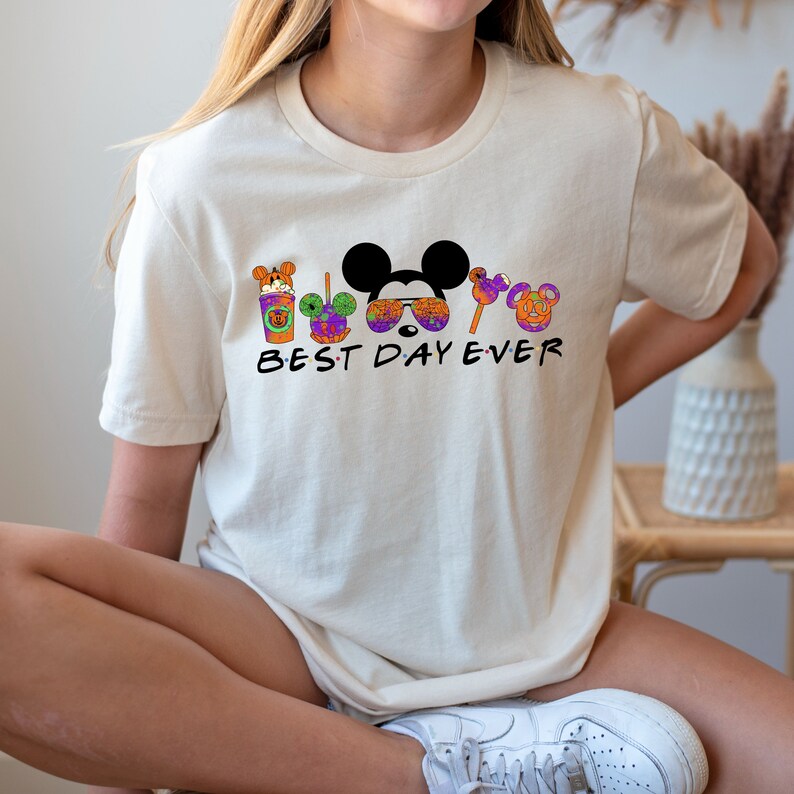 May include: A white t-shirt with a graphic of Mickey Mouse and other Disney characters wearing Halloween costumes. The text "BEST DAY EVER" is printed below the graphic.