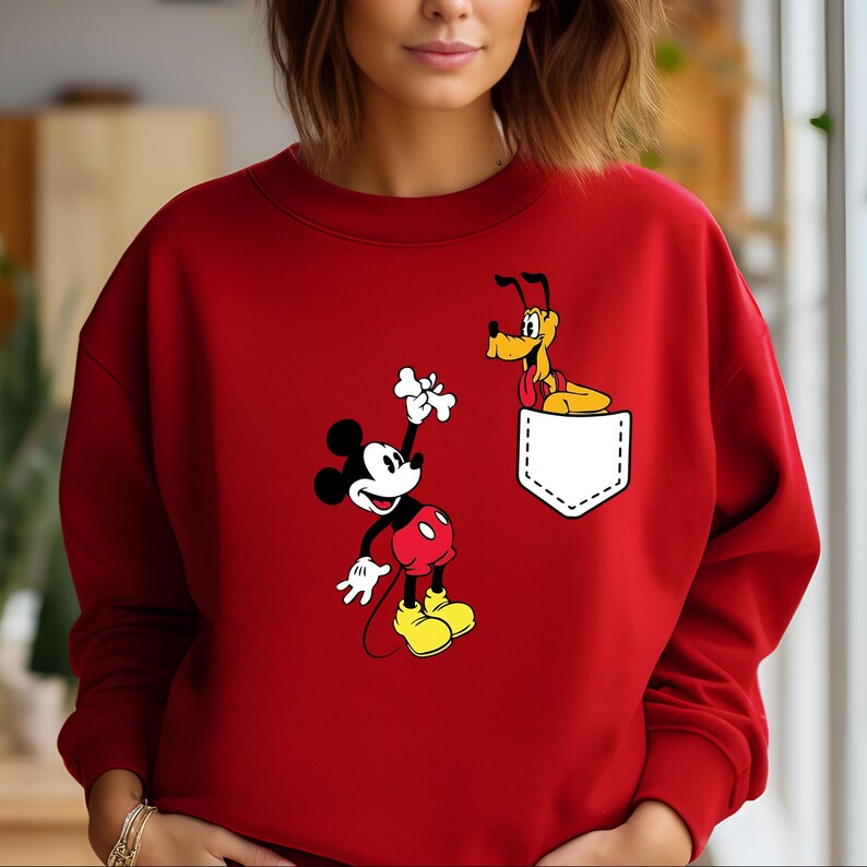 Puede incluir: Sudadera roja con un bolsillo blanco con Mickey Mouse y Pluto. Mickey Mouse sostiene un hueso y Pluto asoma por el bolsillo.