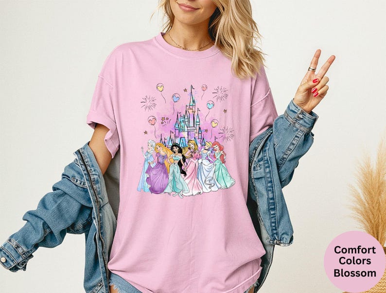 K&ouml;nnte beinhalten: Hellrosa T-Shirt mit einer Grafik eines Schlosses und Prinzessinnen, mit Luftballons und Feuerwerk. Der Text "Comfort Colors Blossom" befindet sich in einem rosa Kreis unten rechts.