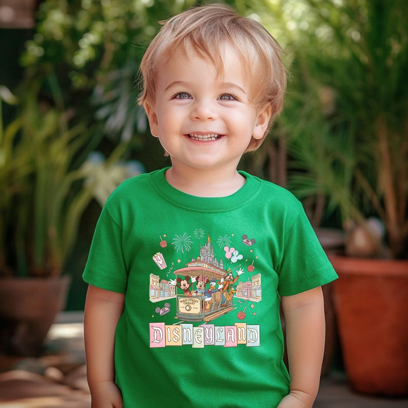 May include: A green t-shirt featuring a Disneyland graphic. The graphic includes a trolley with Mickey Mouse, Donald Duck, and other characters, fireworks, and the word "Disneyland" in a colorful font. The shirt is worn by a young child.
