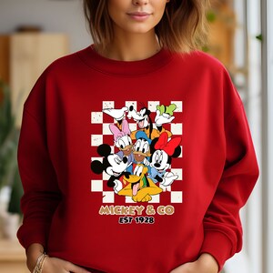 Puede incluir: Sudadera roja con un patr&oacute;n a cuadros con Mickey Mouse y sus amigos. El texto "Mickey & Co. Est 1928" est&aacute; impreso debajo de la imagen.