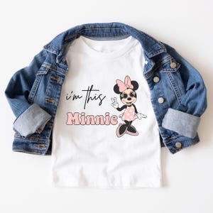 Puede incluir: Camiseta blanca con una caricatura de Minnie Mouse rosa y negra con gafas de sol y un lazo rosa. El texto "i'm this Minnie" est&aacute; impreso en la camiseta en negro.