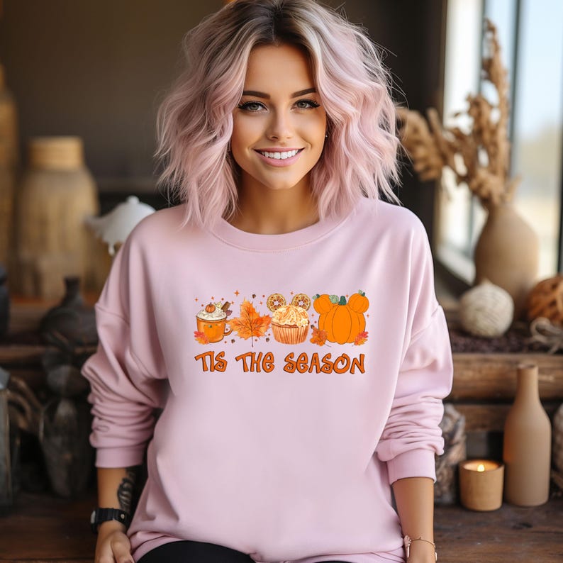 Pu&ograve; includere: Una felpa rosa chiaro a maniche lunghe. La parte anteriore presenta una grafica a tema autunnale con una tazza di caff&egrave;, una foglia d'acero, un cupcake e zucche. Il testo "Tis The Season" &egrave; in basso in arancione.