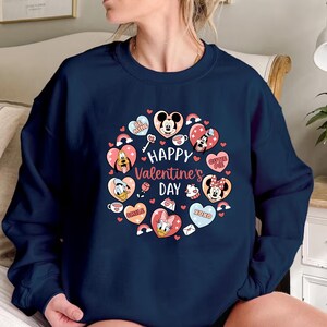 K&ouml;nnte beinhalten: Marineblaues Sweatshirt mit Valentinstagsdesign. Das Design zeigt eine herzf&ouml;rmige Anordnung mit Disney-Figuren, Regenbogen und dem Text "Happy Valentine's Day". Herzen enthalten "Be Mine", "Cutie Pie", "Smile" und "XOXO".