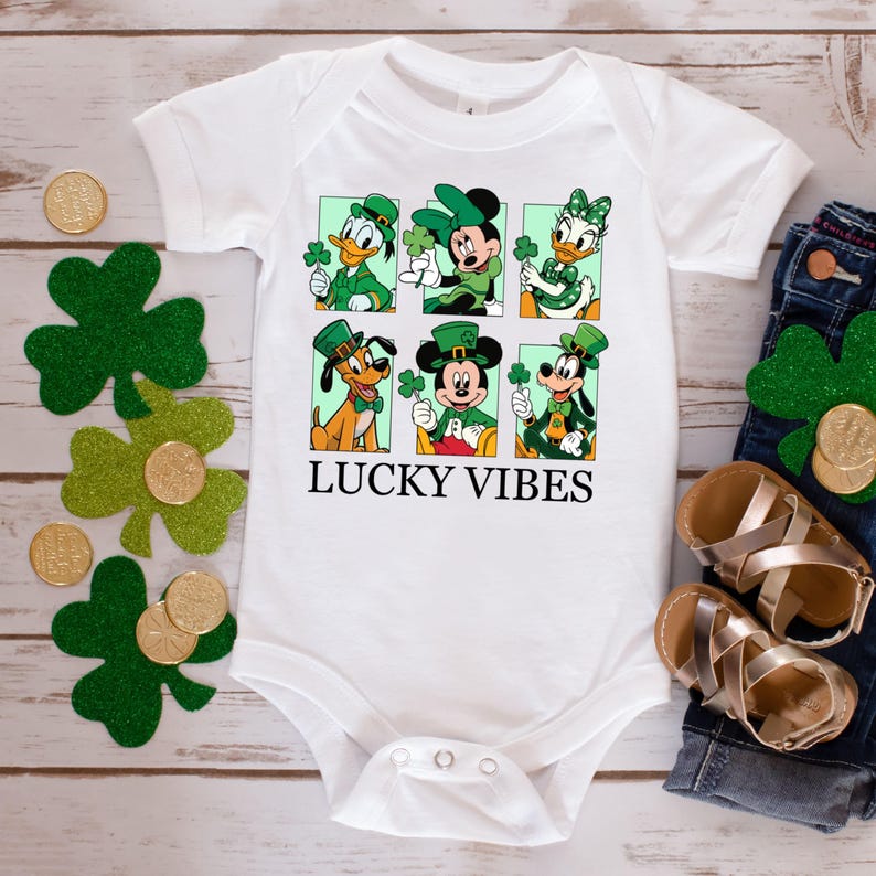 May include: White baby onesie with a green, white, and black graphic of Mickey Mouse, Minnie Mouse, Donald Duck, Goofy, and Pluto wearing green hats and holding shamrocks. The text "LUCKY VIBES" is printed below the graphic.