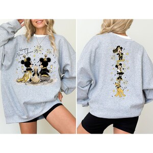 Puede incluir: Sudadera gris claro con Mickey y Minnie Mouse en atuendo formal, celebrando el A&ntilde;o Nuevo. La parte delantera muestra el texto "Happy New Year" con fuegos artificiales. La parte trasera muestra a Mickey, Minnie y Pluto en un dise&ntilde;o festivo.