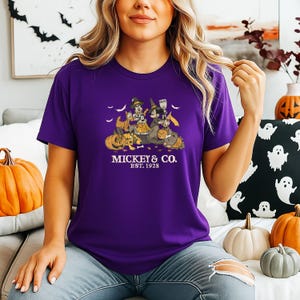 Pode incluir: Uma camiseta roxa com um design de Halloween com Mickey e Minnie Mouse, junto com outros personagens, cercados por ab&oacute;boras e morcegos. O texto "Mickey & Co. Est. 1928" est&aacute; impresso abaixo. A camiseta est&aacute; sendo usada.