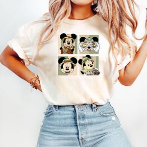 K&ouml;nnte beinhalten: Cremefarbenes T-Shirt mit einem viergeteilten Motiv von Mickey Mouse in Safari-Kleidung. Jedes Feld zeigt Mickey in verschiedenen Posen, mit Safarihut und Outfit, mit Fernglas und Kamera. Das Design ist vor einem Hintergrund aus gr&uuml;nen und beigen Streifen.