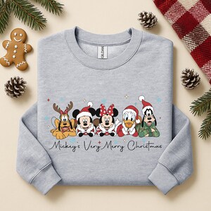 Puede incluir: Una sudadera gris claro con personajes de Disney con atuendos navide&ntilde;os. El dise&ntilde;o incluye a Pluto con cuernos de reno, Mickey Mouse, Minnie Mouse, el Pato Donald y Goofy. El texto "Mickey's Very Merry Christmas" est&aacute; escrito en cursiva.