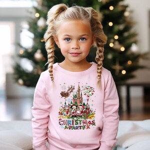 May include: A young girl wearing a light pink sweatshirt with a colorful Christmas Party graphic. The graphic features Disney characters, a castle, Santa's sleigh, and the words "Christmas Party". The background includes a Christmas tree.