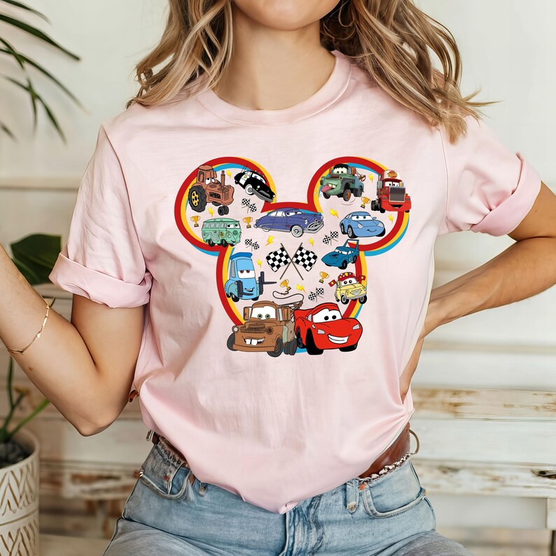 May include: Pink t-shirt with a Mickey Mouse head design featuring various Cars characters, including Mater, Lightning McQueen, and other race cars.