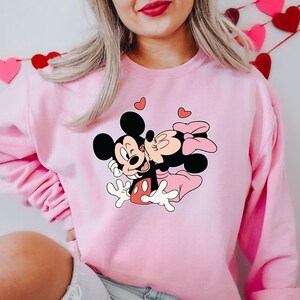 May include: Pink sweatshirt featuring a graphic of Mickey Mouse kissing Minnie Mouse, with hearts floating above them. The cartoon characters are in black, white, red, and pink. The sweatshirt is a solid light pink color.