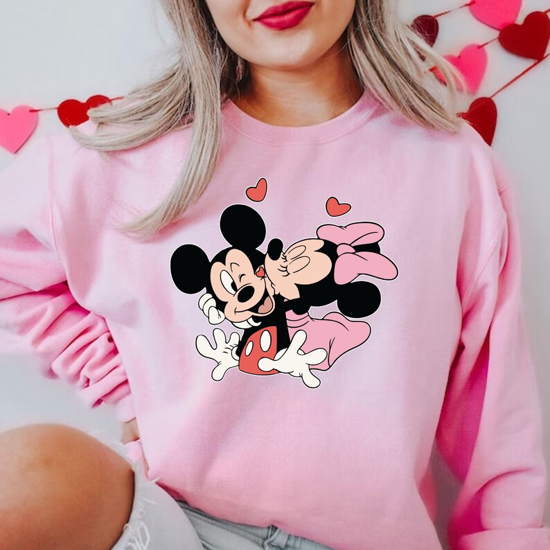 May include: Pink sweatshirt featuring a graphic of Mickey Mouse kissing Minnie Mouse, with hearts floating above them. The cartoon characters are in black, white, red, and pink. The sweatshirt is a solid light pink color.