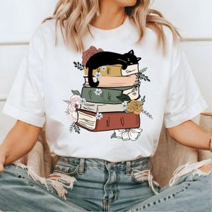 May include: White t-shirt with a black cat sleeping on a stack of books. The books are decorated with flowers and have a vintage aesthetic.