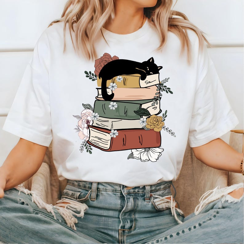 May include: White t-shirt with a black cat sleeping on a stack of books. The books are decorated with flowers and have a vintage aesthetic.