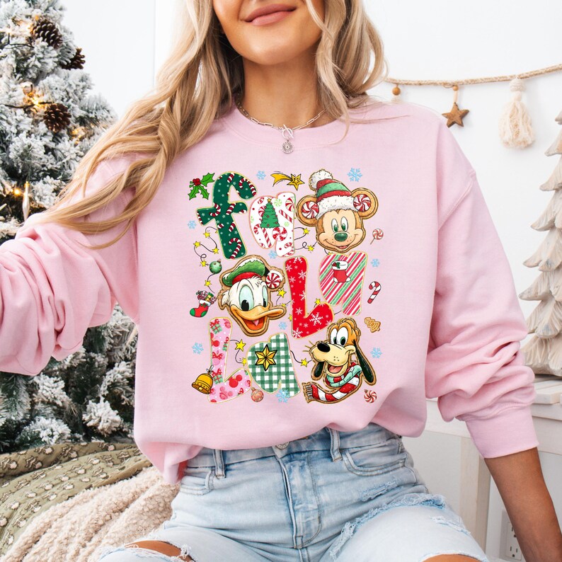 K&ouml;nnte beinhalten: Ein hellrosa Sweatshirt mit dem Wort "FALL" in einem festlichen Design. Die Buchstaben sind mit Weihnachtselementen wie Stechpalmen, Zuckerstangen und Schneeflocken verziert. Micky Maus, Donald Duck und Pluto sind ebenfalls abgebildet.