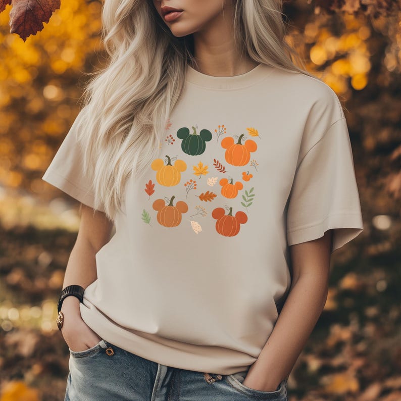 May include: Beige t-shirt featuring a colorful pumpkin design with Mickey Mouse ears and autumn leaves. The pumpkins are in shades of orange, yellow, and green. The shirt has short sleeves and a crew neck.