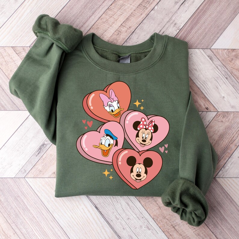 Puede incluir: Sudadera verde con un gr&aacute;fico de Mickey Mouse, Minnie Mouse, Donald Duck y Daisy Duck en corazones rosas.
