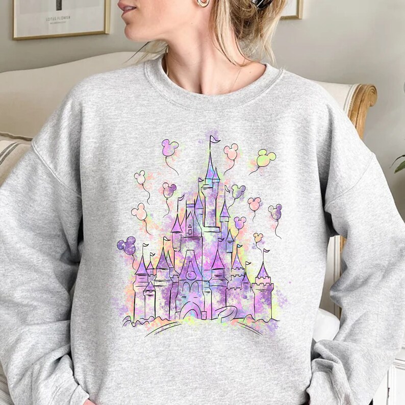 May include: A grey sweatshirt featuring a watercolour illustration of a castle with Mickey Mouse ears floating around it.
