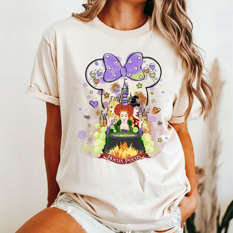 May include: A white t-shirt with a purple and white Minnie Mouse head design featuring the Sanderson sisters from the movie Hocus Pocus. The design includes a castle, fireworks, and the text "Hocus Pocus".