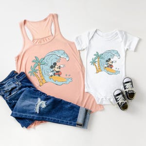 May include: A pink tank top and a white bodysuit featuring a graphic of Mickey Mouse surfing on a wave. The graphic also includes palm trees.