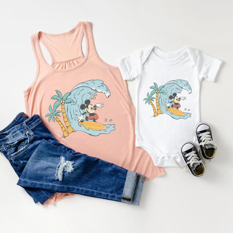 May include: A pink tank top and a white bodysuit featuring a graphic of Mickey Mouse surfing on a wave. The graphic also includes palm trees.