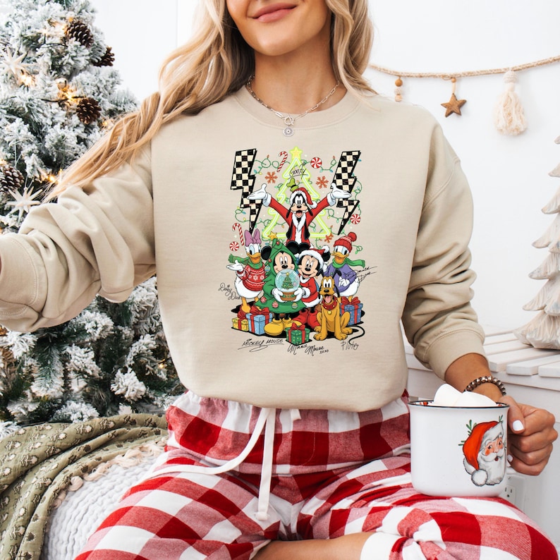 May include: Beige crewneck sweatshirt featuring a festive graphic of Disney characters, including Mickey Mouse, Donald Duck, and Goofy, celebrating Christmas. The design includes a Christmas tree, presents, and candy canes. The sweatshirt has a relaxed fit.
