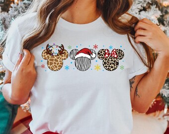 May include: White t-shirt with a Christmas-themed design featuring three animal-themed Mickey Mouse ears. The ears are decorated with a reindeer, zebra, and leopard print.