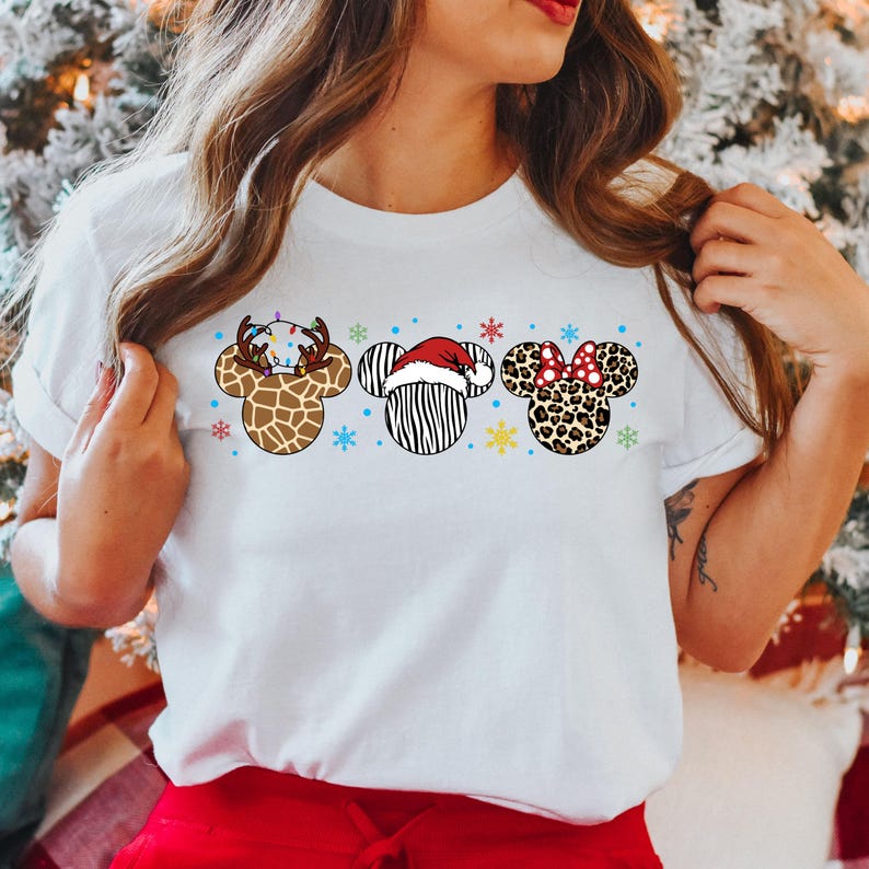 May include: White t-shirt with a Christmas-themed design featuring three animal-themed Mickey Mouse ears. The ears are decorated with a reindeer, zebra, and leopard print.