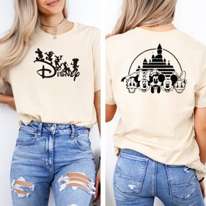 Puede incluir: Una camiseta beige con un logotipo de Disney negro y personajes en la parte delantera y una silueta negra del castillo de Disney con Mickey Mouse y sus amigos en la parte posterior.