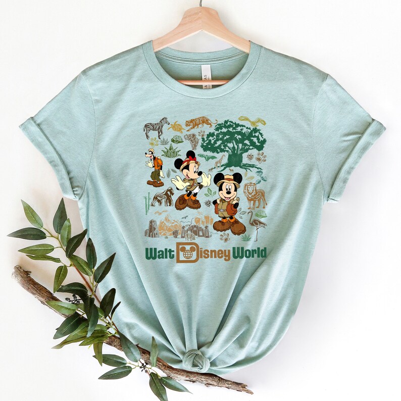 May include: A light blue t-shirt featuring a graphic print of Mickey Mouse and Goofy dressed in safari outfits. The design also includes a range of animals and plants, such as a zebra, a lion, a giraffe, and a tree. The text "Walt Disney World" is printed beneath the graphic.