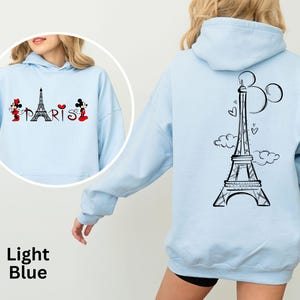 K&ouml;nnte beinhalten: Hellblauer Kapuzenpullover mit schwarzem Eiffelturm-Motiv und dem Wort "PARIS" auf der Vorderseite. Auf der R&uuml;ckseite befindet sich ein gr&ouml;&szlig;erer Eiffelturm mit Mickey Mouse Ohren. Der Hoodie ist hellblau.
