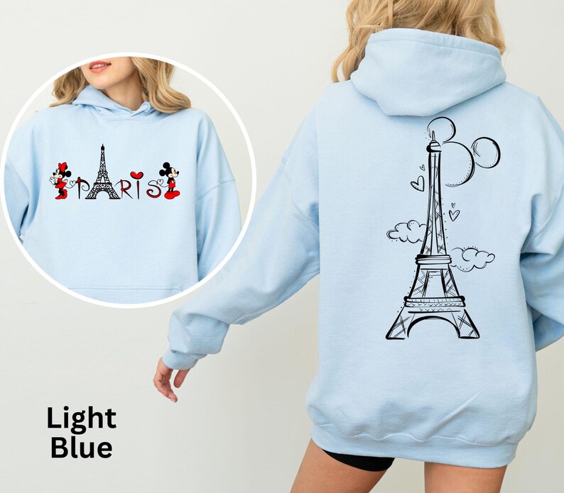 K&ouml;nnte beinhalten: Hellblauer Kapuzenpullover mit schwarzem Eiffelturm-Motiv und dem Wort "PARIS" auf der Vorderseite. Auf der R&uuml;ckseite befindet sich ein gr&ouml;&szlig;erer Eiffelturm mit Mickey Mouse Ohren. Der Hoodie ist hellblau.